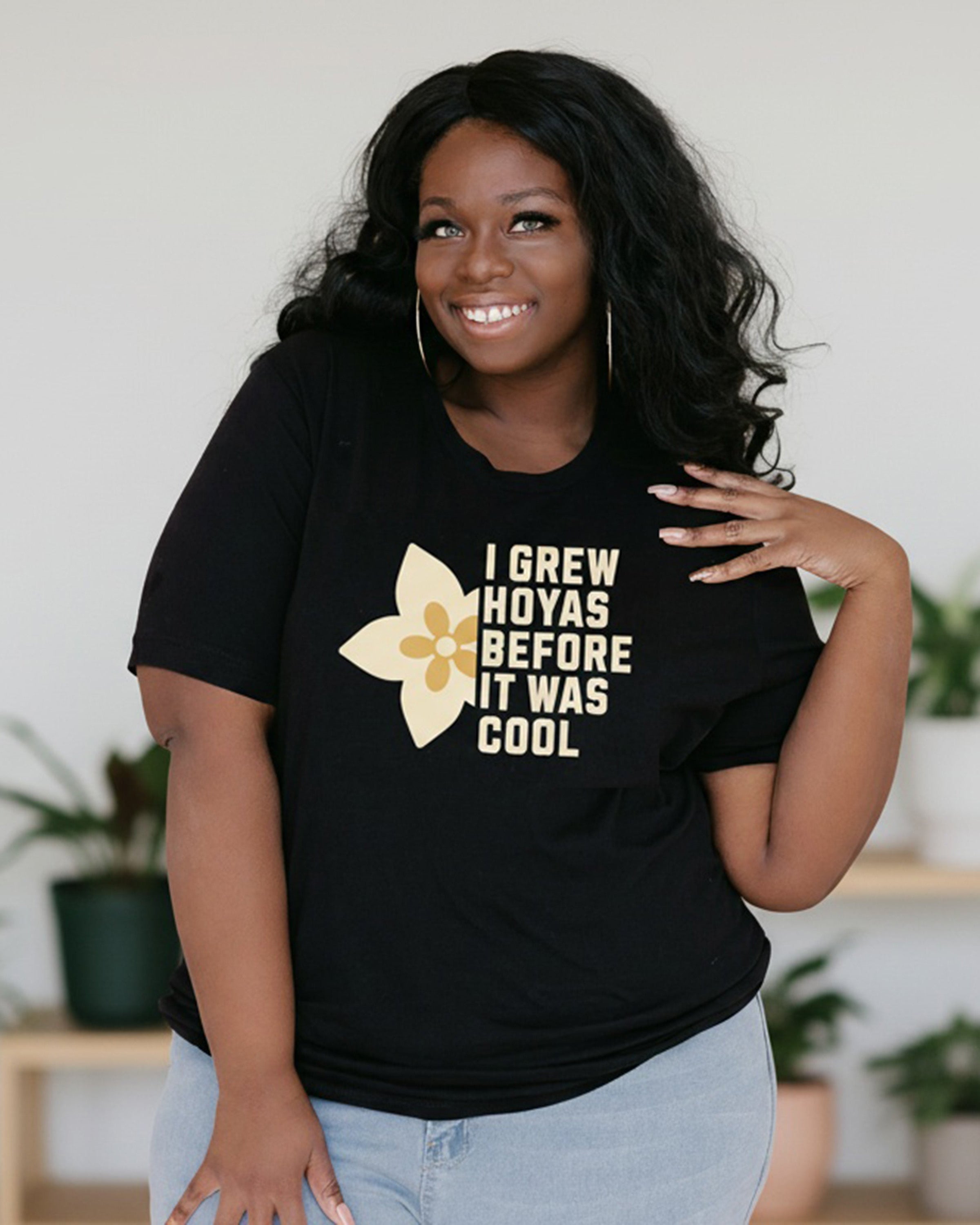 Woman wearing a black t-shirt with a graphic and text, standing in a room with plants.
