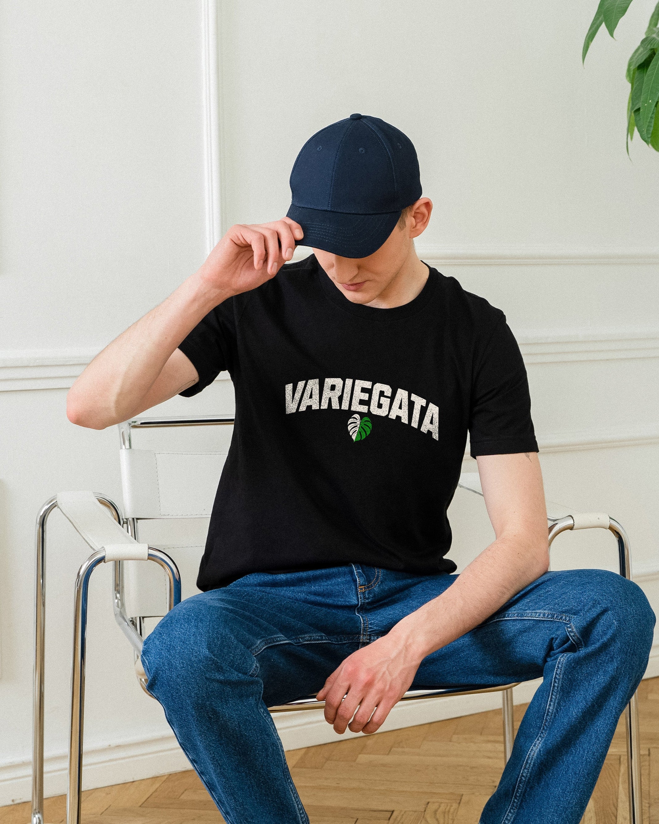 Person wearing a black t-shirt with 'VARIEGATA' text sitting in a room with a plant and table.
