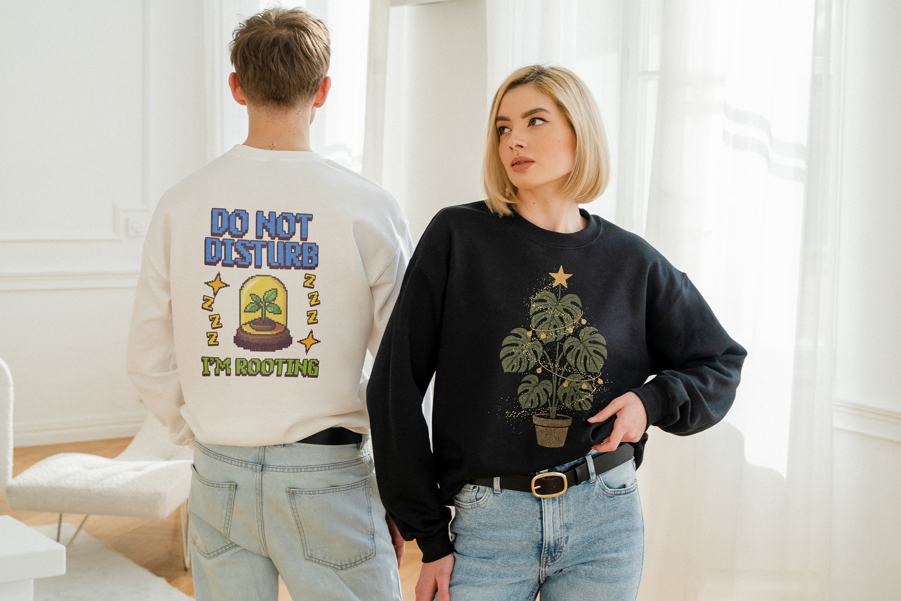 Two models indoors wearing plant-themed t-shirts. Woman in white shirt reads “Today’s good mood sponsored by Blooms,” man in olive tee with skull graphic says “Dead Plant Club.” Styled with black shorts and beige pants, standing by a large potted plant.
