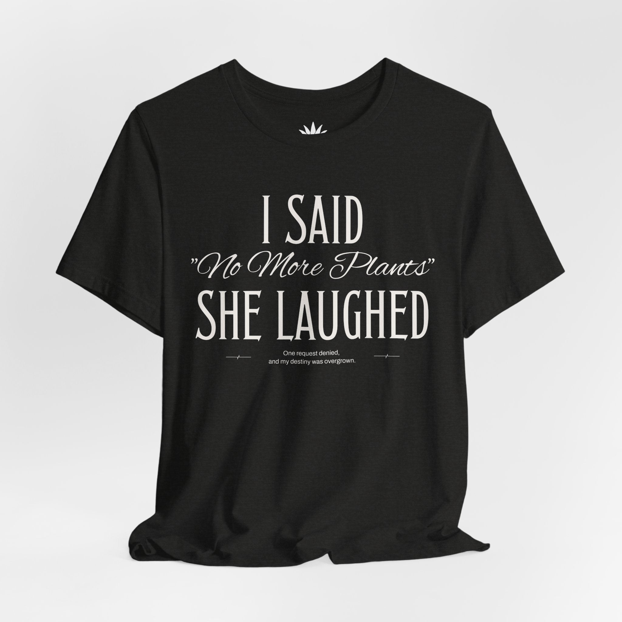 Black t-shirt with white vintage lettering that reads “I Said ‘No More Plants’ She Laughed,” a funny graphic tee for plant lovers and collectors. Hanging.